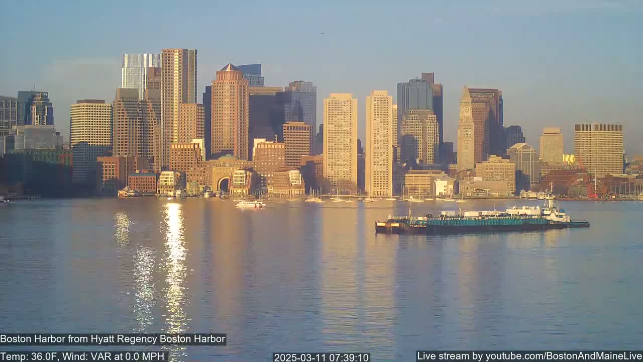 A calm harbor reflects the sunlit cityscape of Boston, with a barge in the foreground.