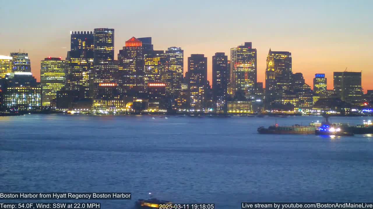 A brightly lit city skyline sits across a dark body of water at dusk, with a barge visible in the foreground and a strong south-southwesterly wind.