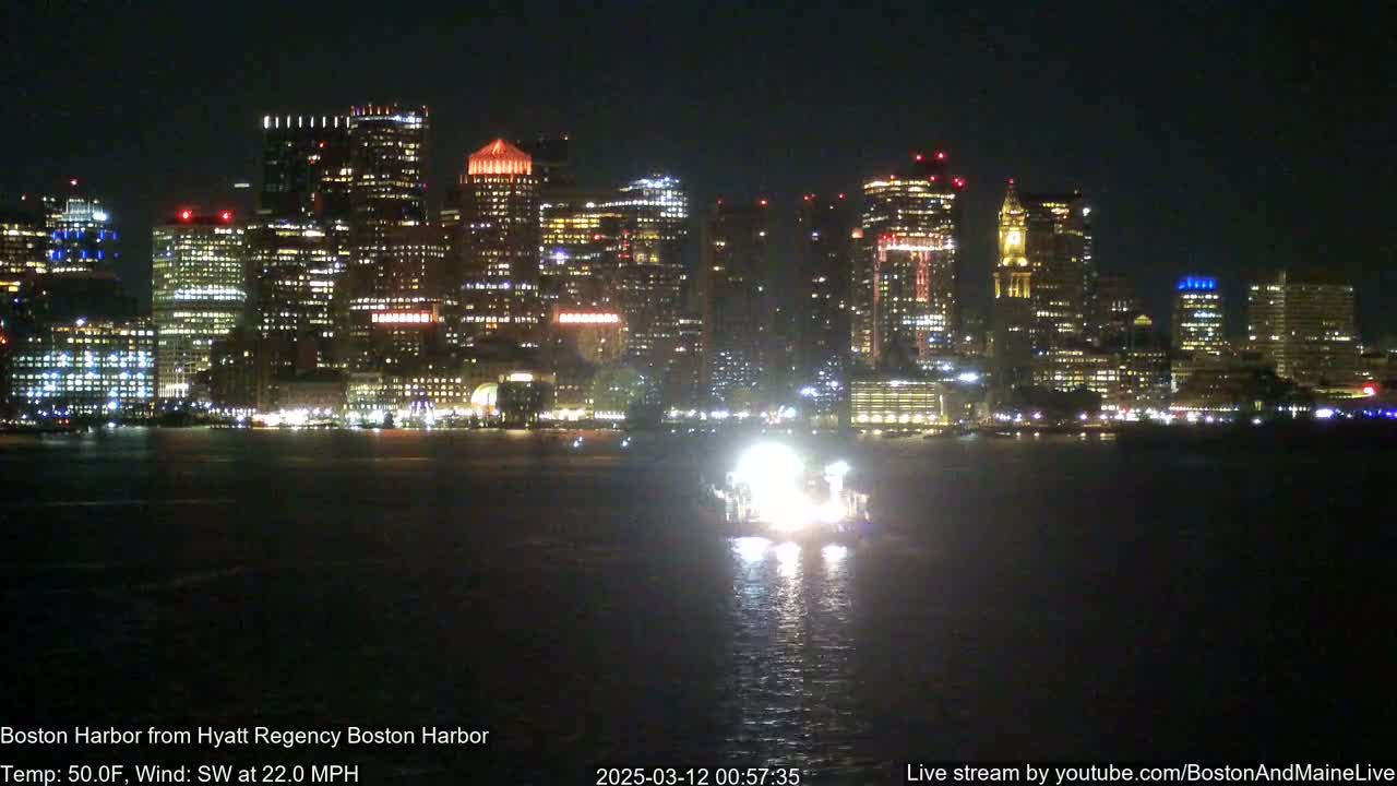 A brightly lit city skyline is seen across a dark body of water at night, with a boat in the foreground and a southwest wind at 22 mph.