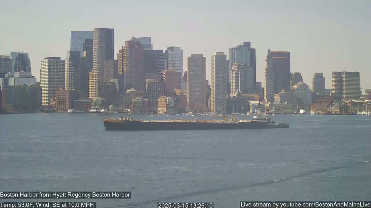 A large tanker sits in calm water in front of a city skyline under a hazy sky.