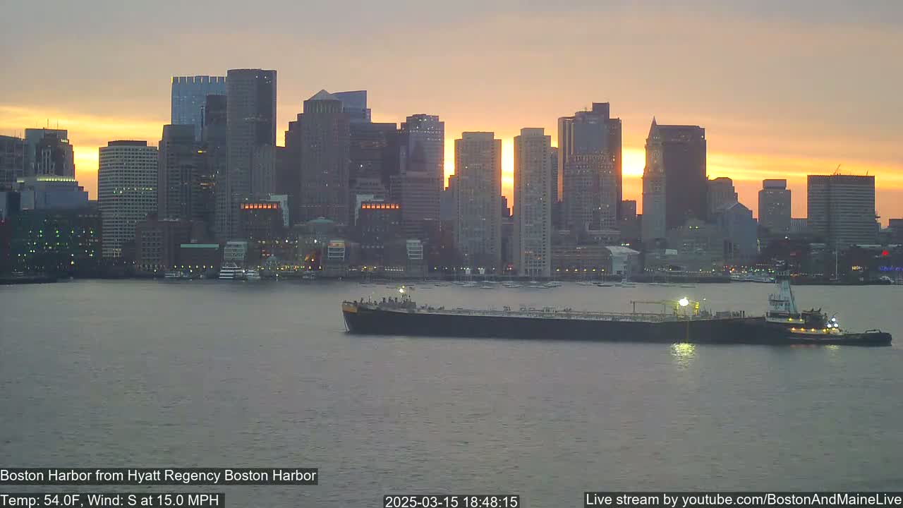 A large tanker and tugboat are in the harbor at sunset, with a city skyline in the background under a mostly clear sky.