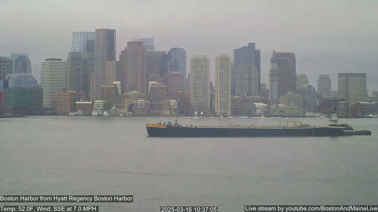 A barge is being towed across a harbor in front of a city skyline on an overcast day with a SSE wind at 7.0 mph and a temperature of 52°F.