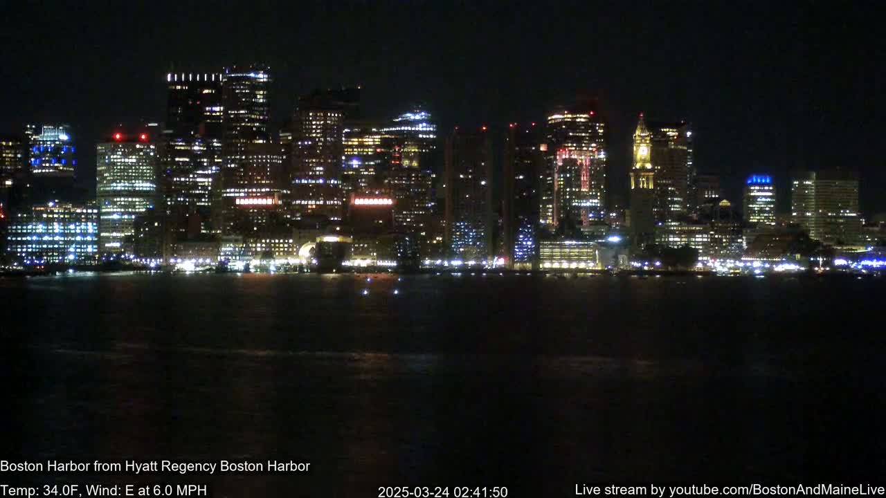 A brightly lit city skyline is visible across a dark body of water at night, under a clear sky with a 34°F temperature and a 6.0 mph east wind.