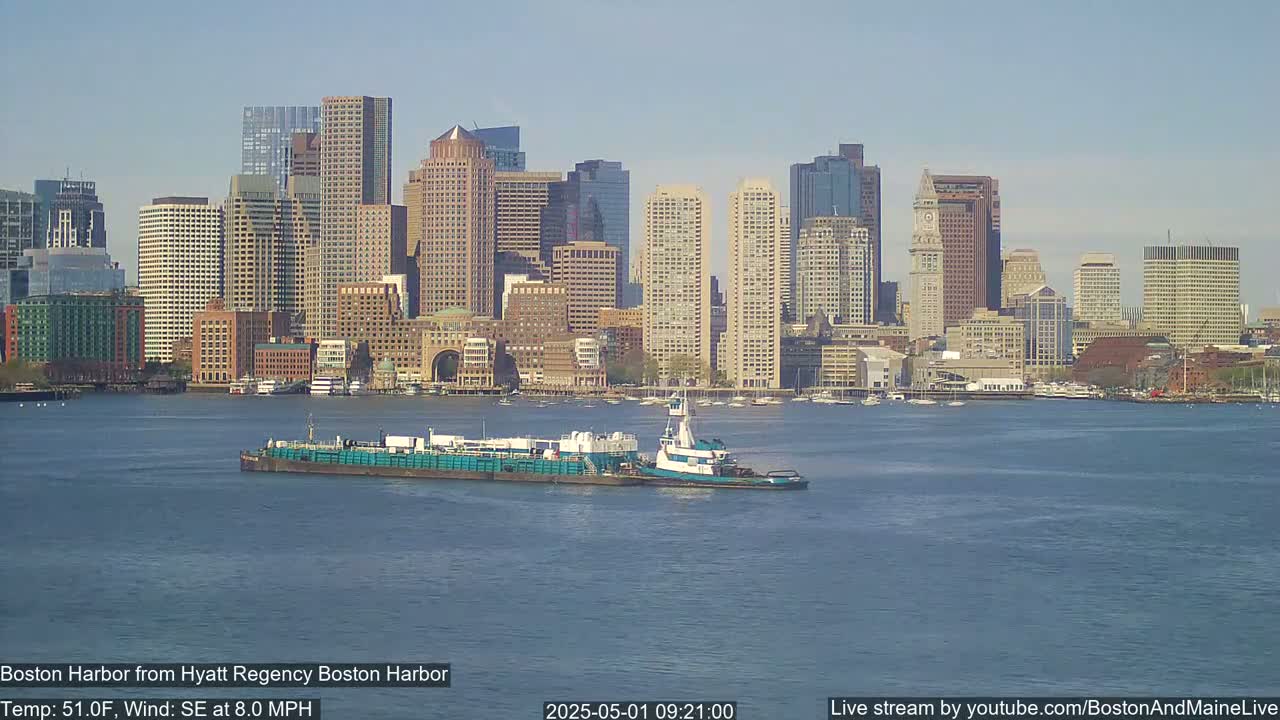 A tugboat pushes a barge across a calm harbor in front of a city skyline under a clear sky.
