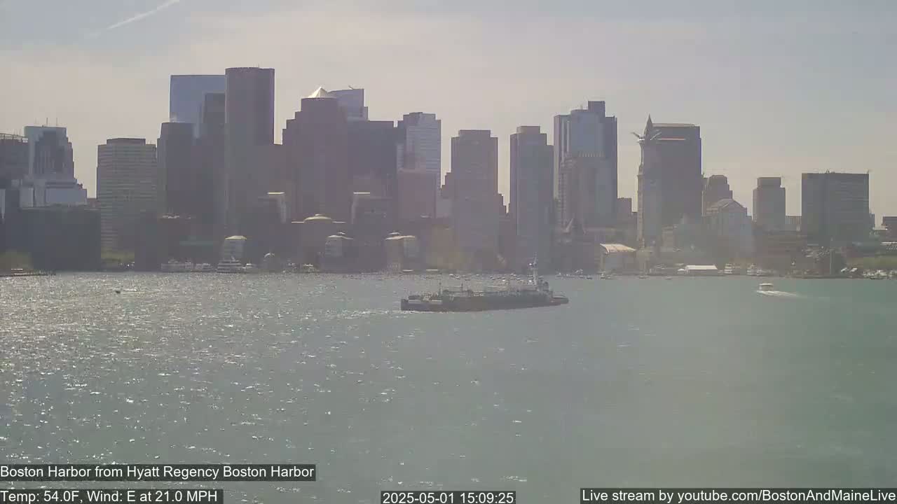 A tugboat moves across a harbor in front of a hazy city skyline on a partly sunny day with an east wind.