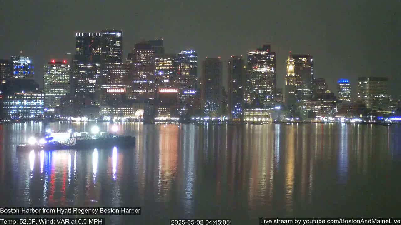 A nighttime view of a city skyline reflected in calm water, with a vessel visible in the foreground.