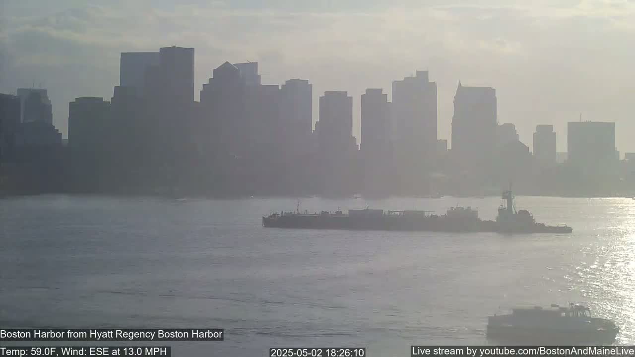 A hazy cityscape is visible across a body of water where a tugboat pushes a barge; a slight chop is visible on the water's surface.