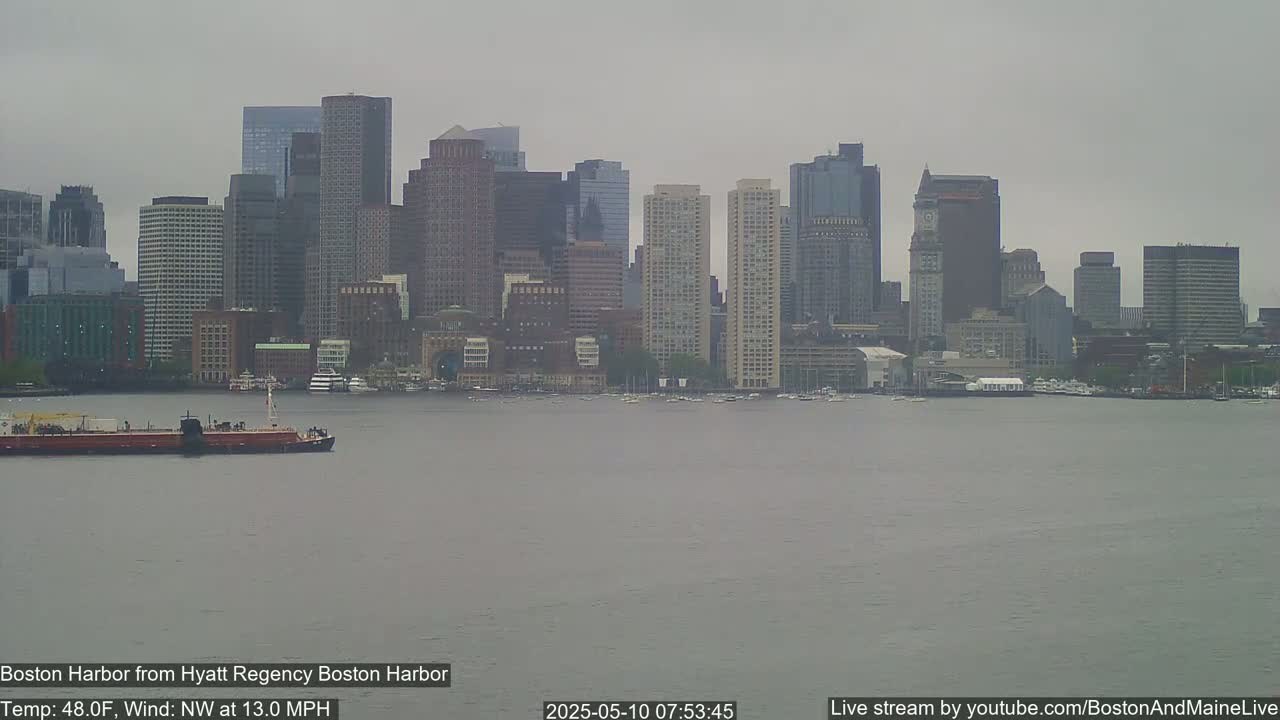 A barge floats on a gray, calm harbor in front of a city skyline under an overcast sky.