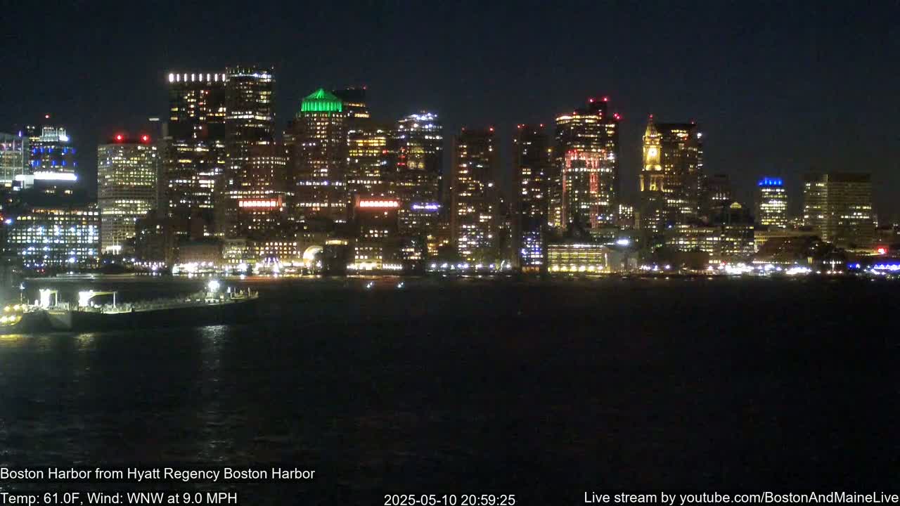 A brightly lit cityscape is seen across a dark body of water at night, under a clear sky with a 9 mph wind.