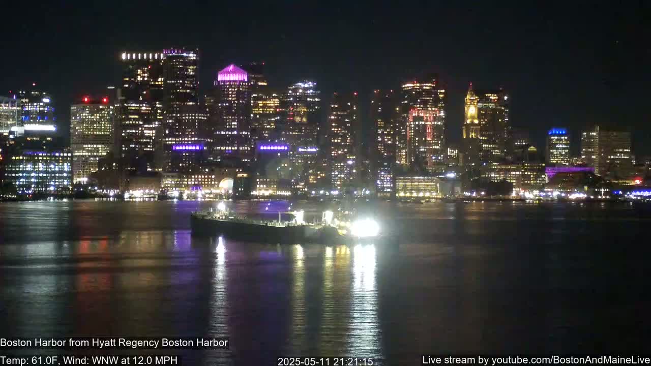 A brightly lit city skyline is reflected in calm harbor water at night, with a large vessel in the foreground.