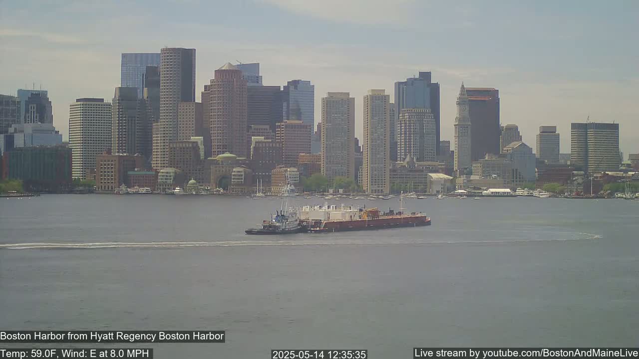 A tugboat pushes a barge across a calm harbor in front of a city skyline on a partly cloudy day with an east wind at 8 mph and a temperature of 59°F.