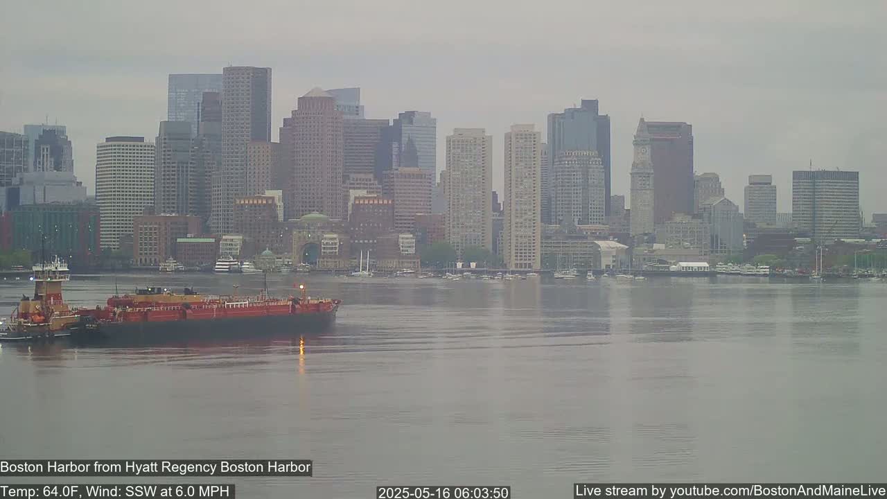 A tugboat pushes a barge across a hazy harbor in front of a cityscape on a cloudy day.