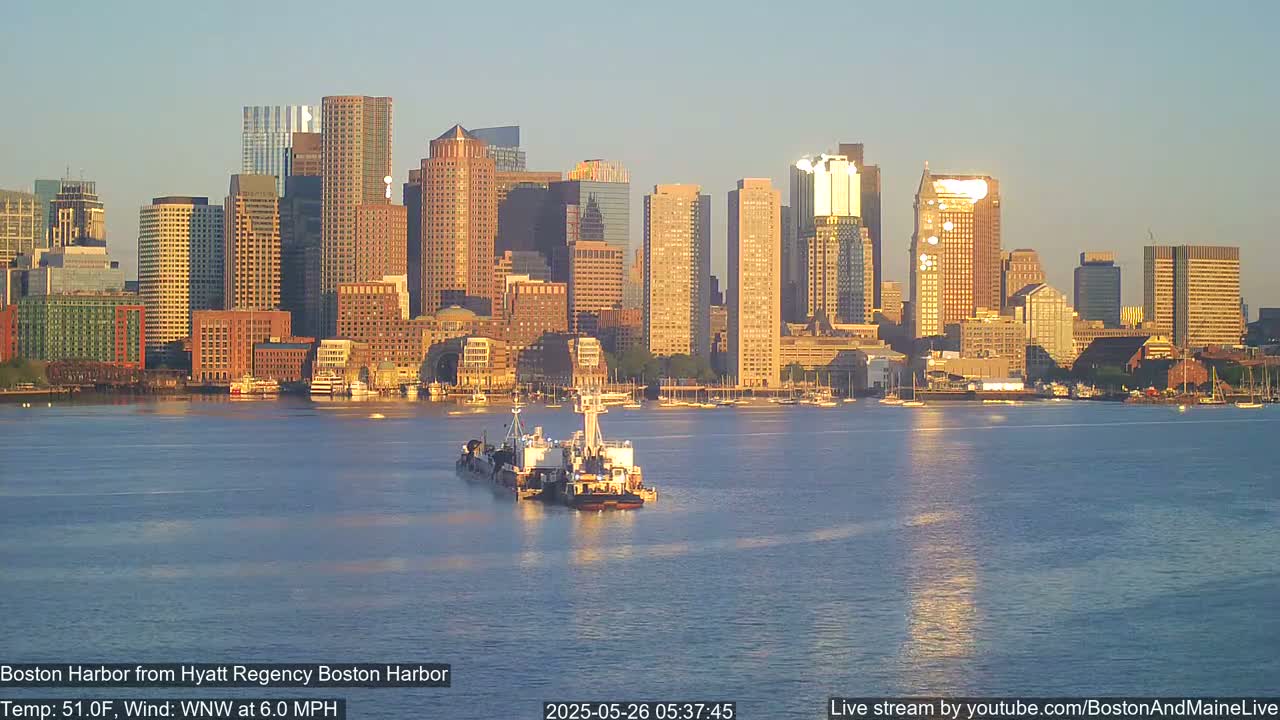 A tugboat pushes a barge on calm water in front of a city skyline at sunrise, with a gentle westerly wind.