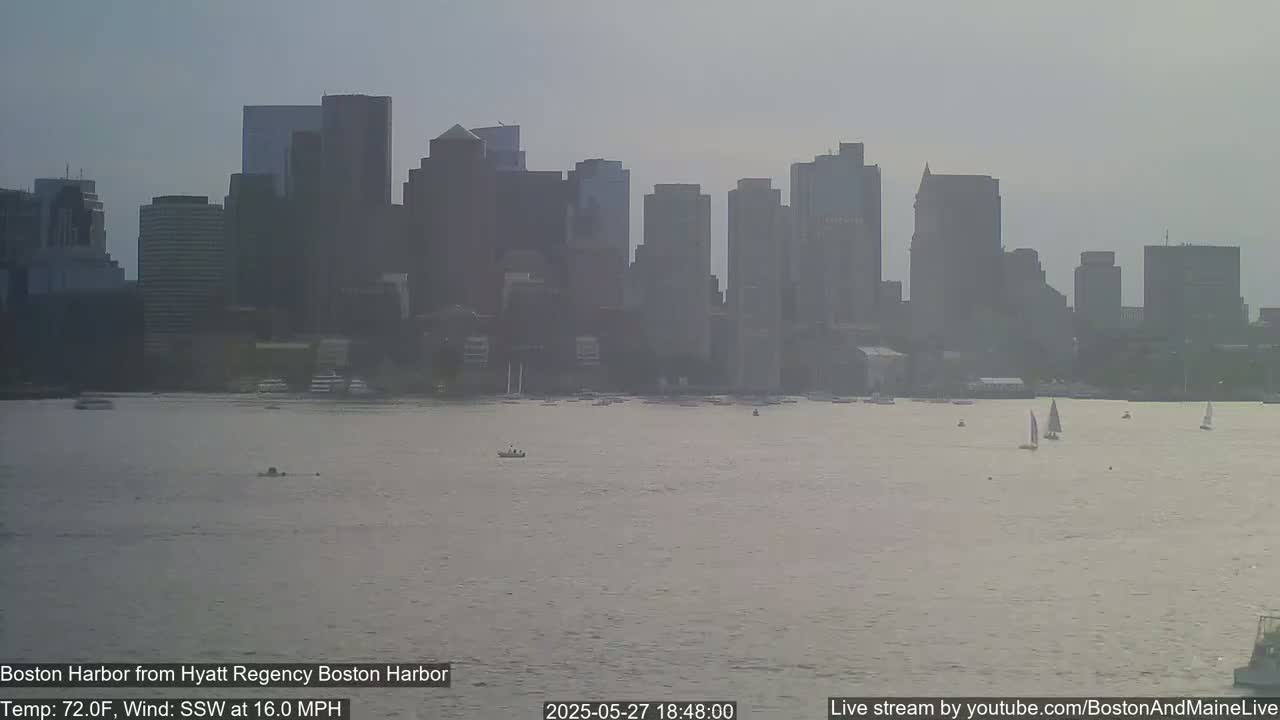 A hazy view of the Boston skyline across a calm harbor, with a few sailboats and small boats visible on the water under a light, hazy sky.