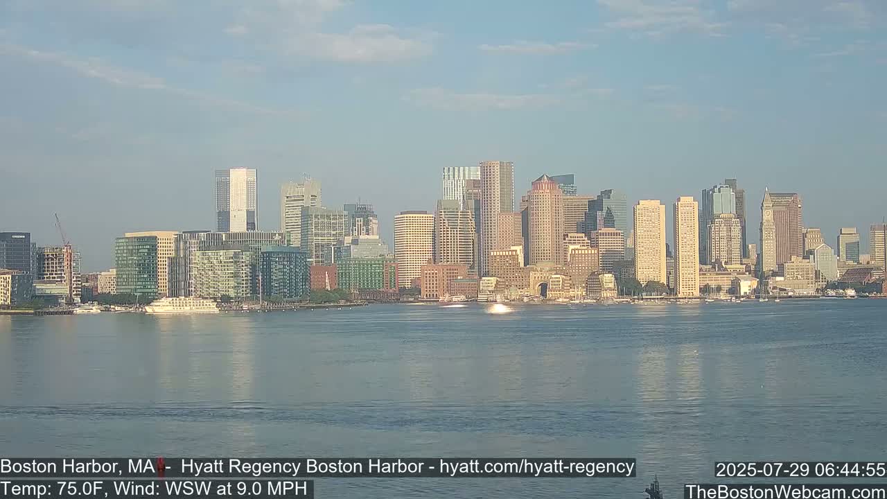 A hazy, sunny day shows a city skyline across a calm body of water with a few small boats.
