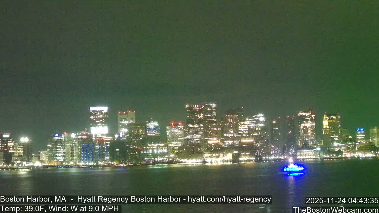 A brightly lit city skyline stretches across a calm harbor at night, featuring a boat with blue lights on the water, under clear skies with a temperature of 39.0F and a west wind at 9.0 MPH.