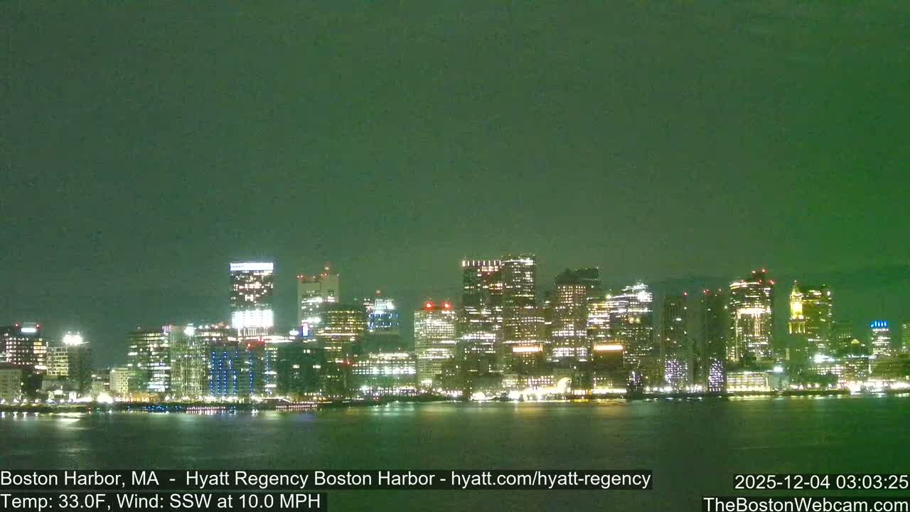 A brightly lit city skyline is seen at night across a dark body of water under a green-tinted sky, with the temperature at 33.0F and wind SSW at 10.0 MPH.