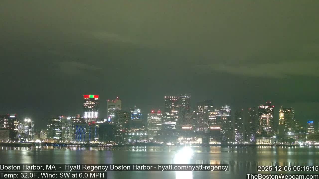 A nighttime view of the Boston city skyline, brightly lit with numerous buildings, reflects upon the dark harbor water under an overcast sky and cold temperatures.