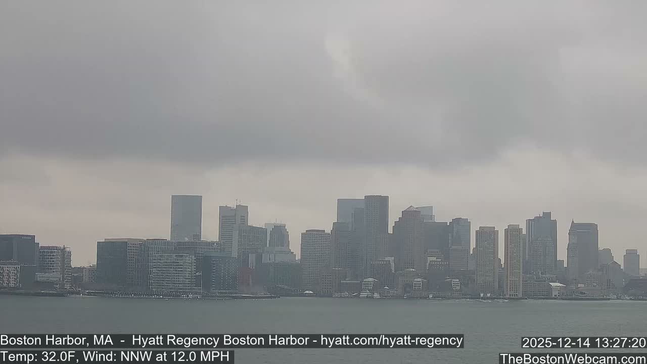 A nighttime view of the Boston city skyline, brightly lit with numerous buildings, reflects upon the dark harbor water under an overcast sky and cold temperatures.