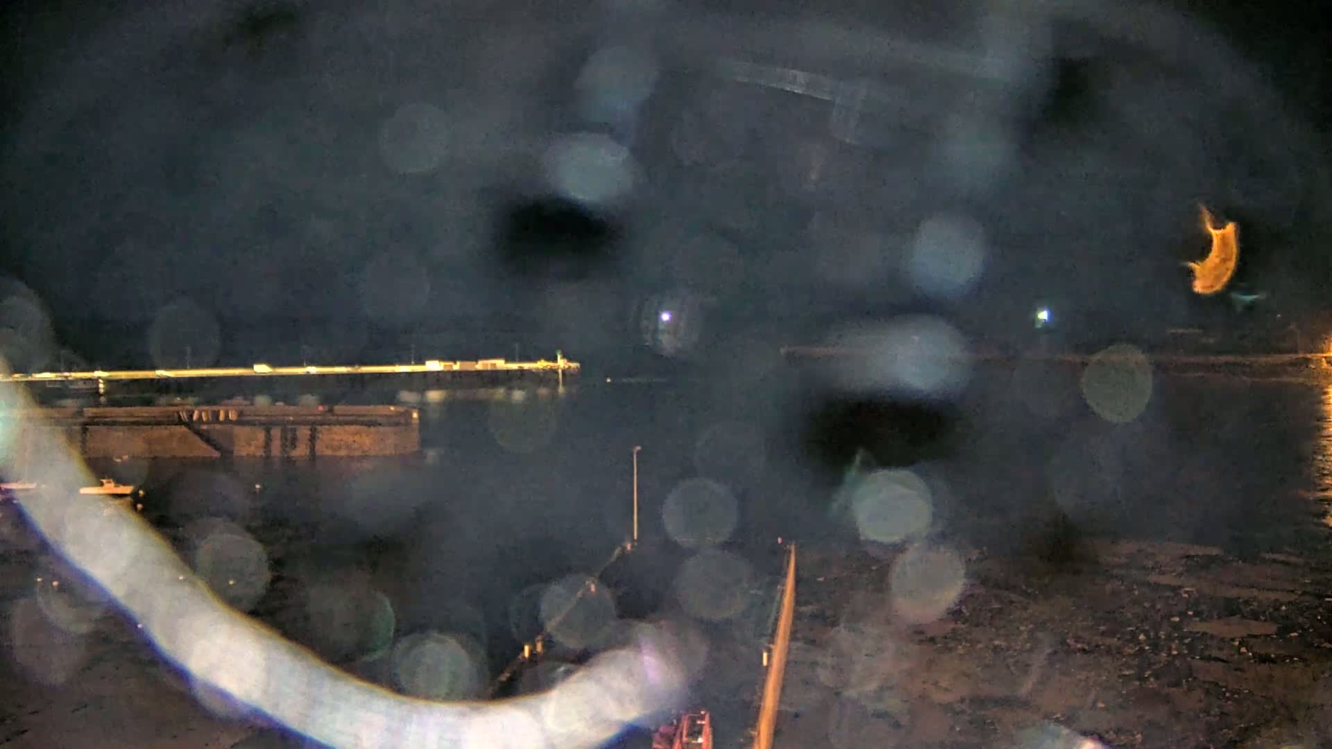 A nighttime view of a harbor with lights visible across the water, seen through rain-covered glass.