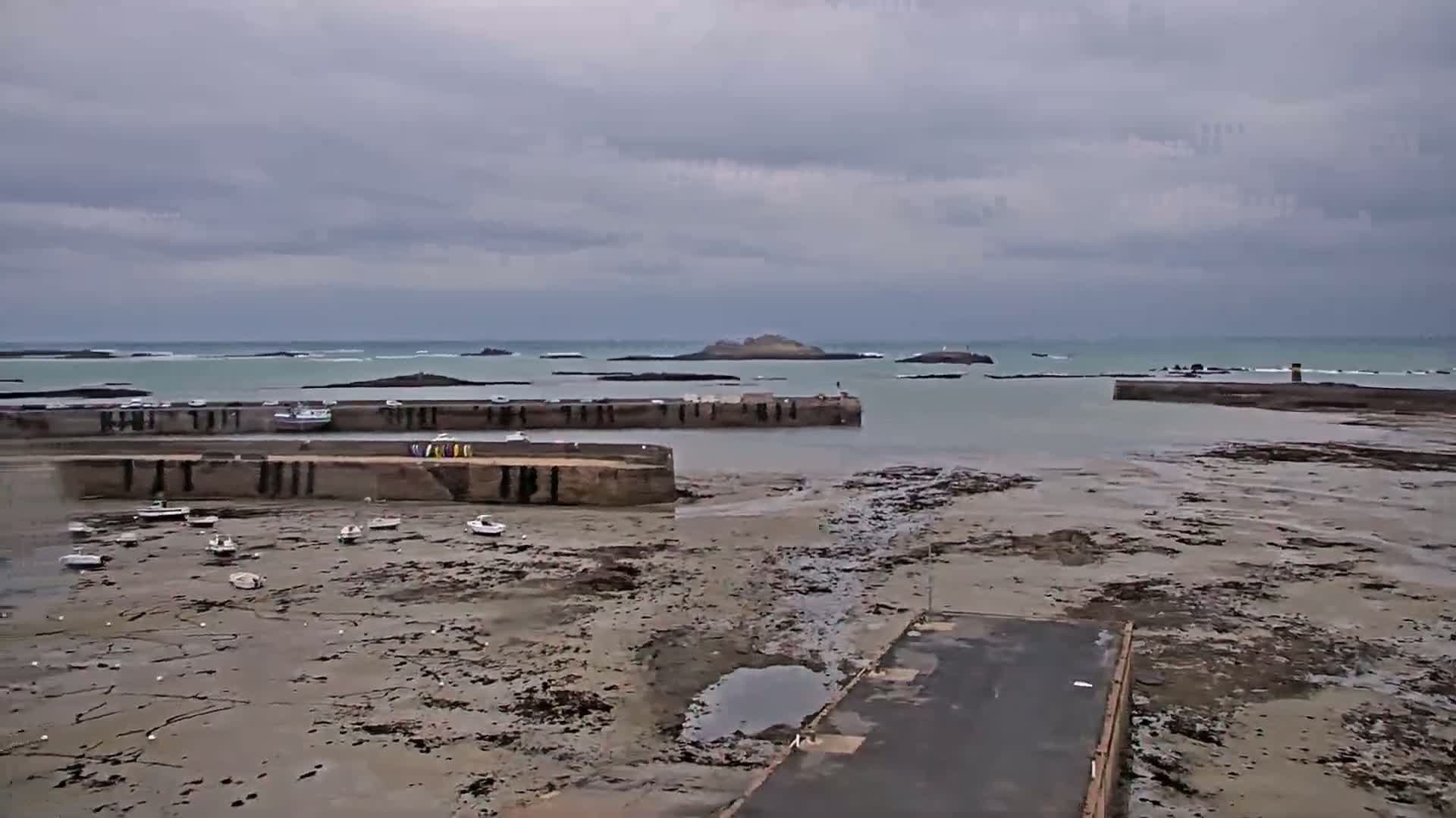 Under an overcast sky, a harbor filled with small boats sits exposed at low tide, with rocky islands visible in the distance.