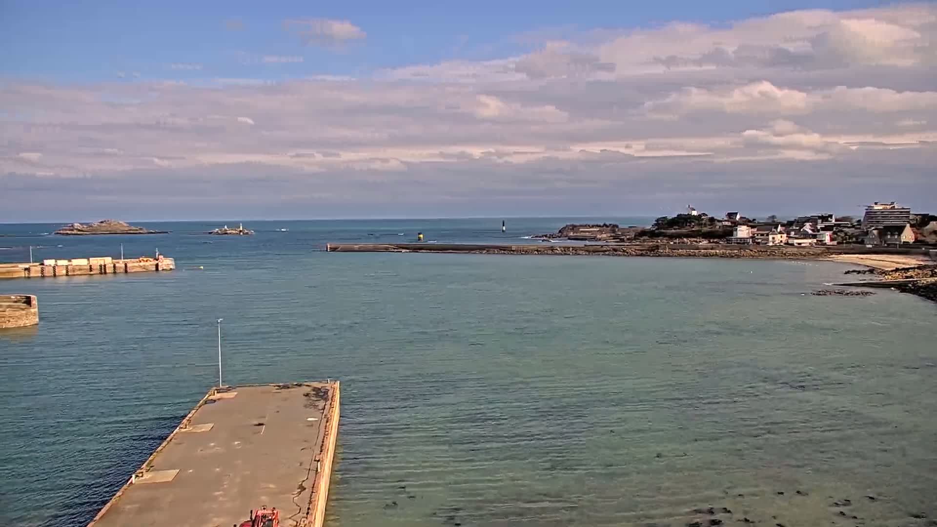 A partly cloudy day reveals a coastal scene with a harbor, breakwater, small town, and islands in the distance.