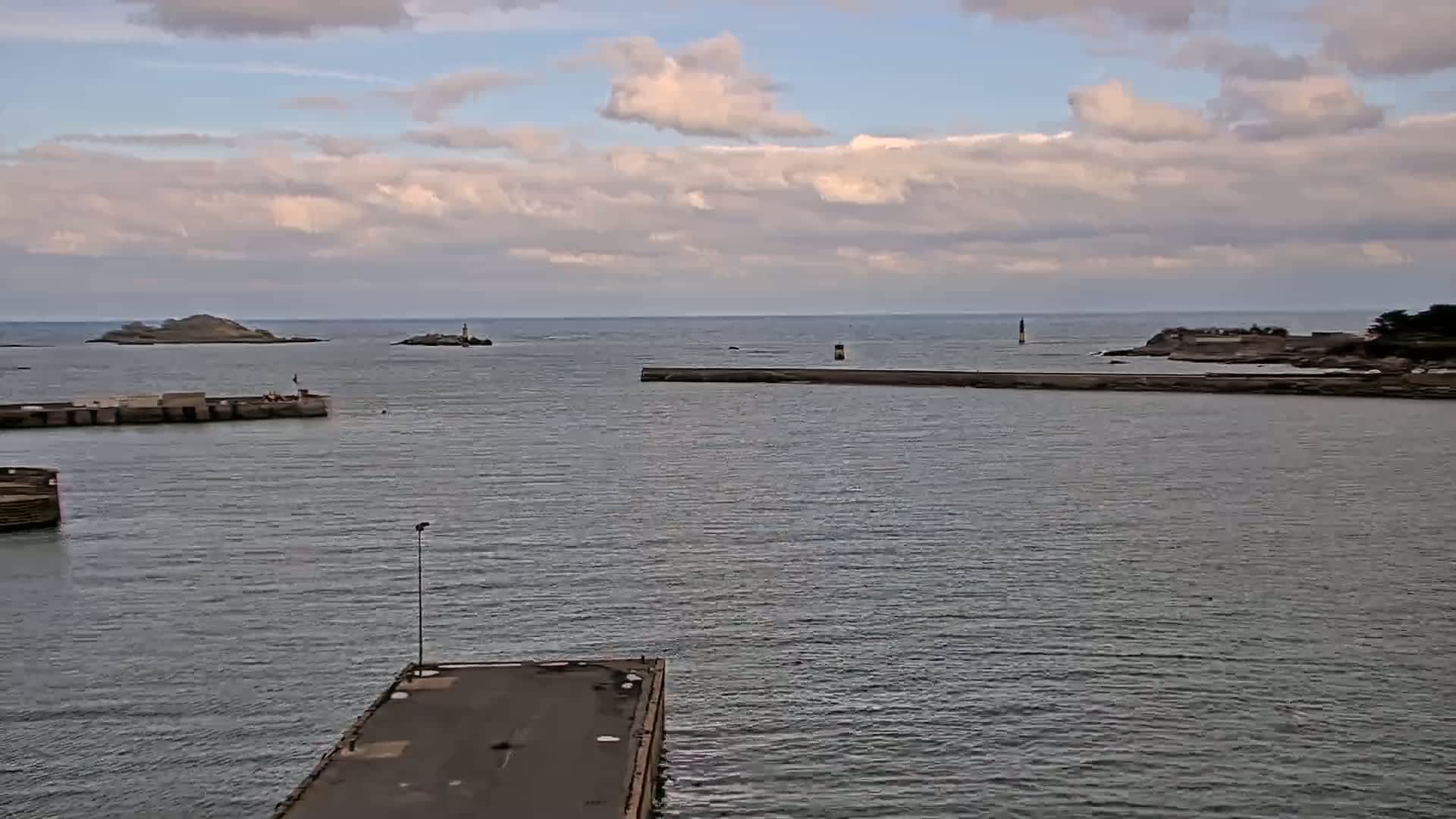 A harbor with calm water under a partly cloudy sky shows small islands and a breakwater in the distance.