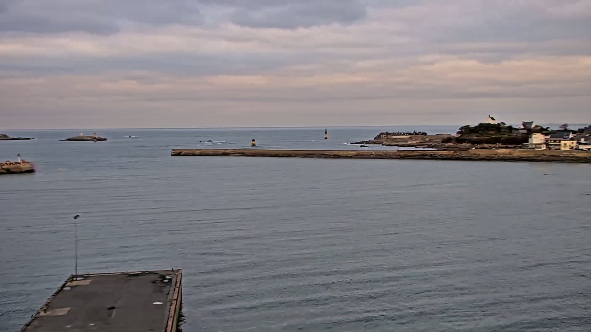 A calm, overcast day reveals a coastal scene with a harbor, breakwater, small islands, and a distant village nestled on a rocky shoreline.