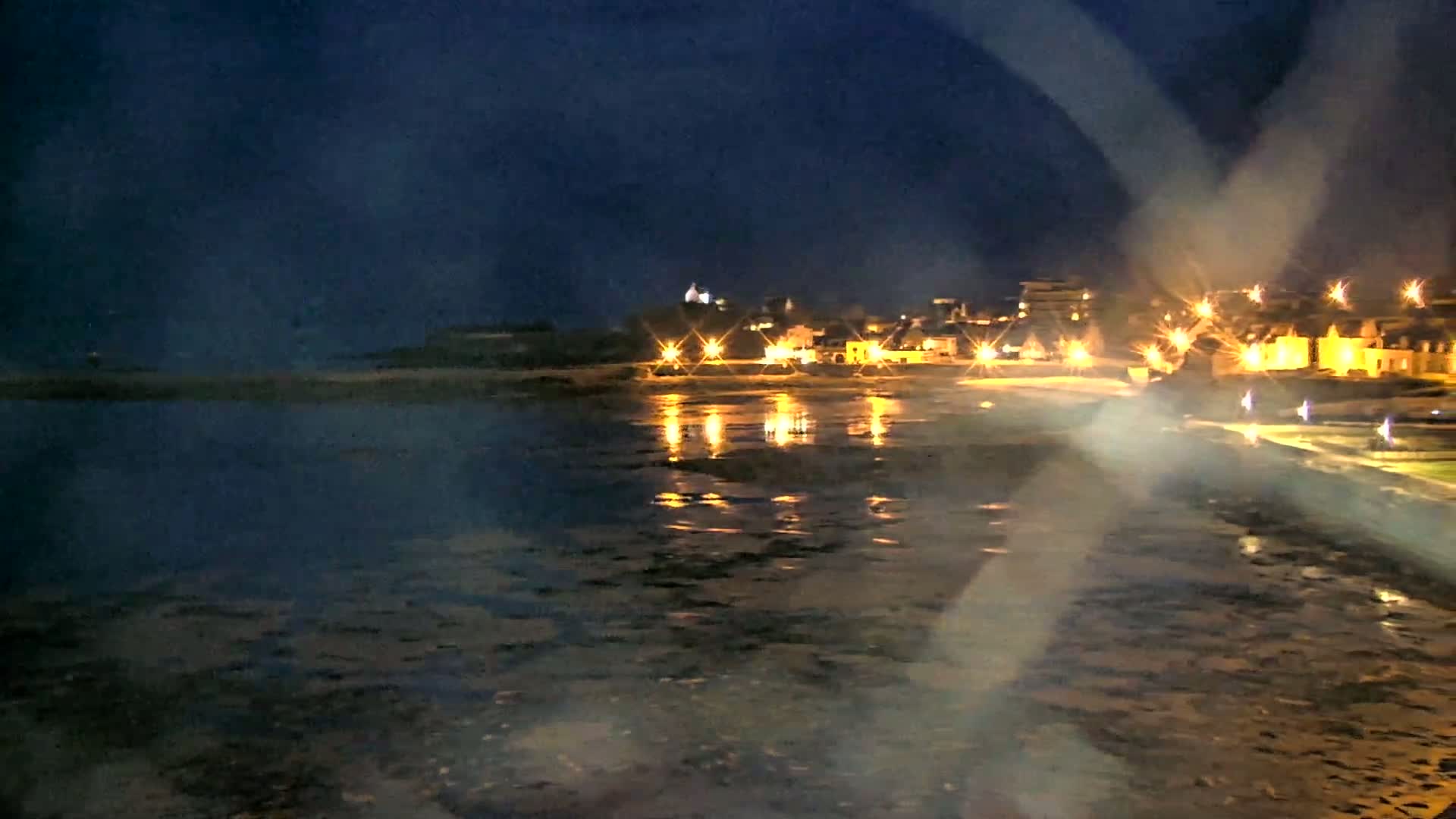 A nighttime view of a coastal town with its lights reflecting on a dark, shallow body of water under a dark sky.