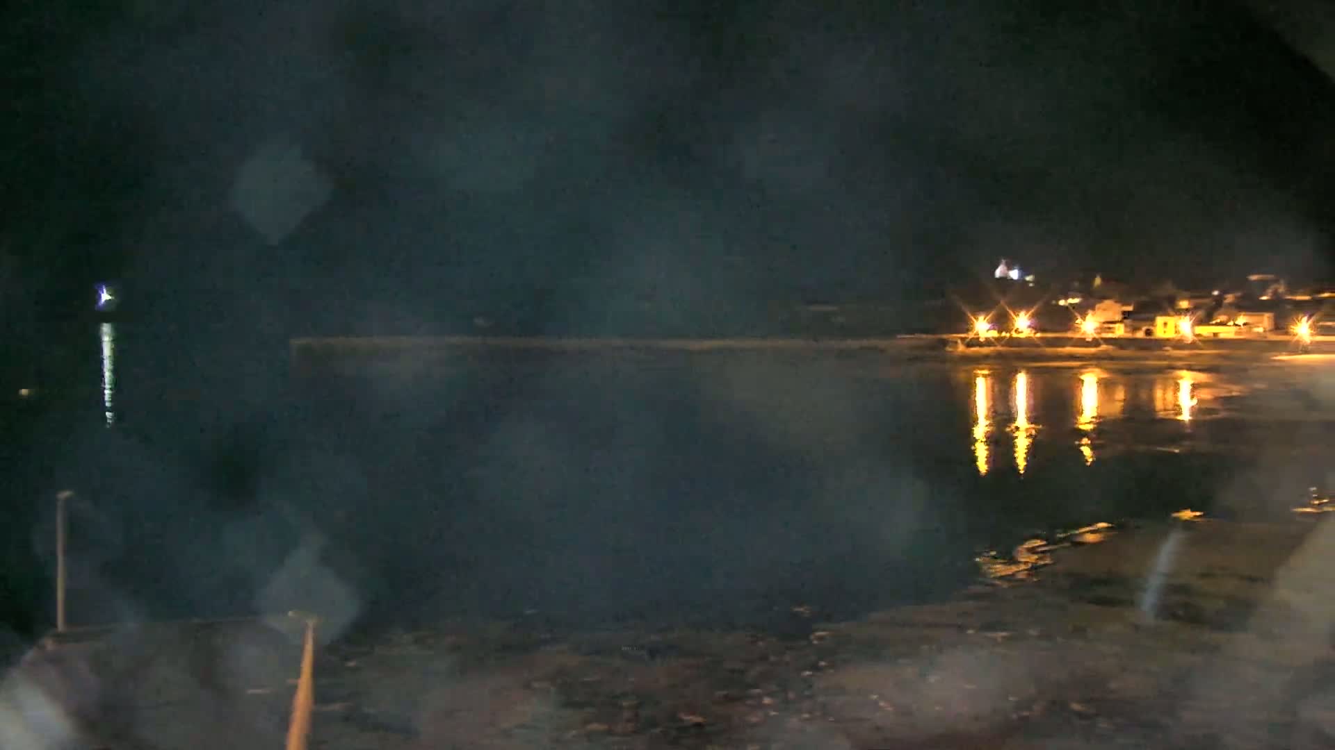 A nighttime view of a calm body of water reflecting the lights of a small town across the shore, under hazy conditions.
