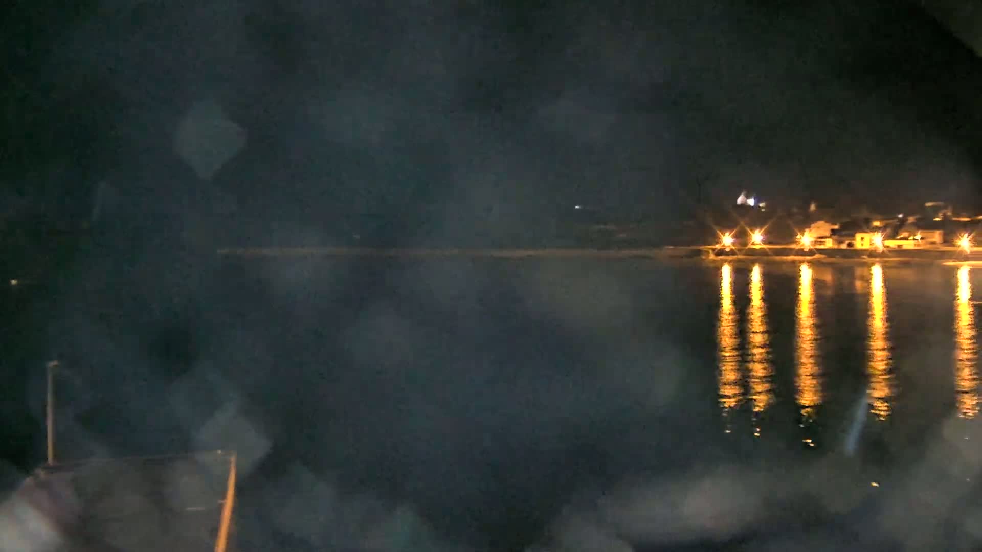 A night-time view of a calm body of water reflecting the lights of a distant, small town, seen through a hazy, possibly rain-streaked, window.
