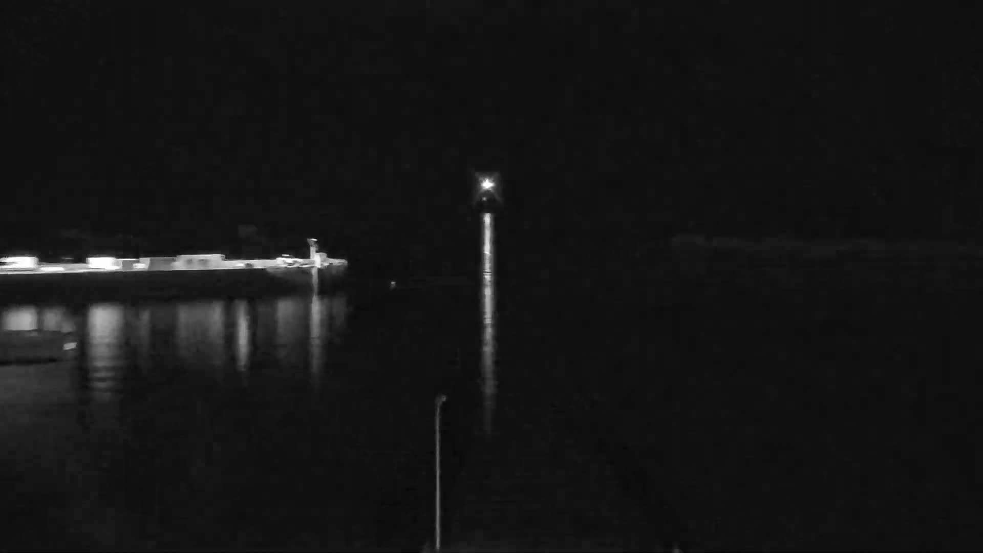 A nighttime view shows a long, bright reflection on dark water extending from a light source in the distance, with a dimly lit structure visible on the left bank.