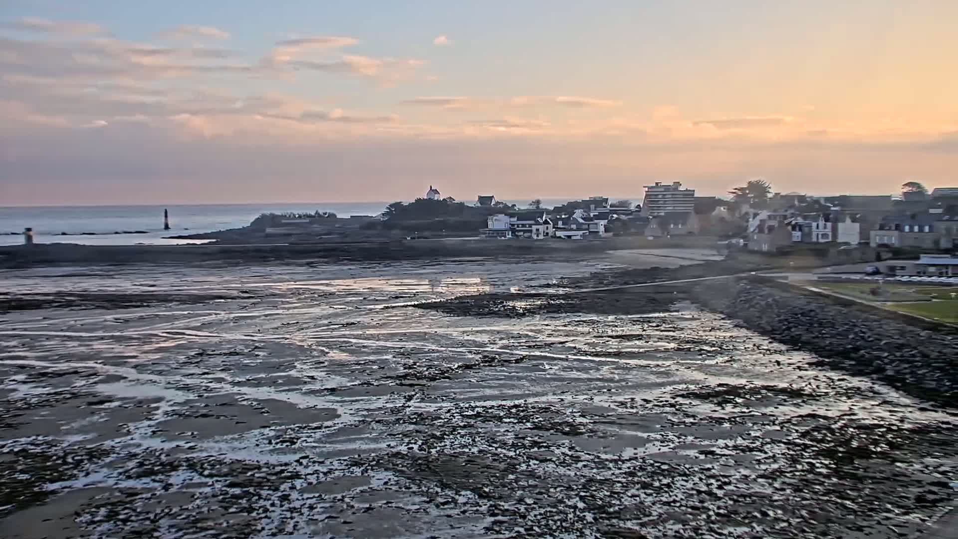 A coastal town with a lighthouse in the distance is seen at low tide under a partly cloudy, sunset sky.
