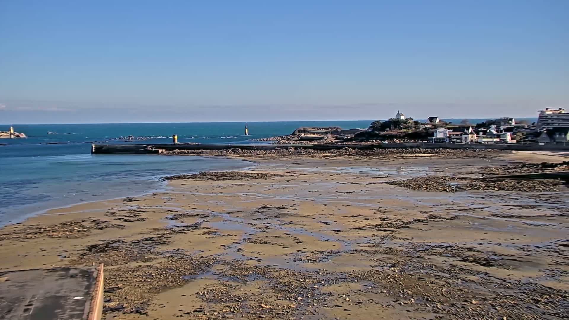 A coastal town is visible across a wide expanse of exposed, sandy seabed with small pools of water, under a clear blue sky.