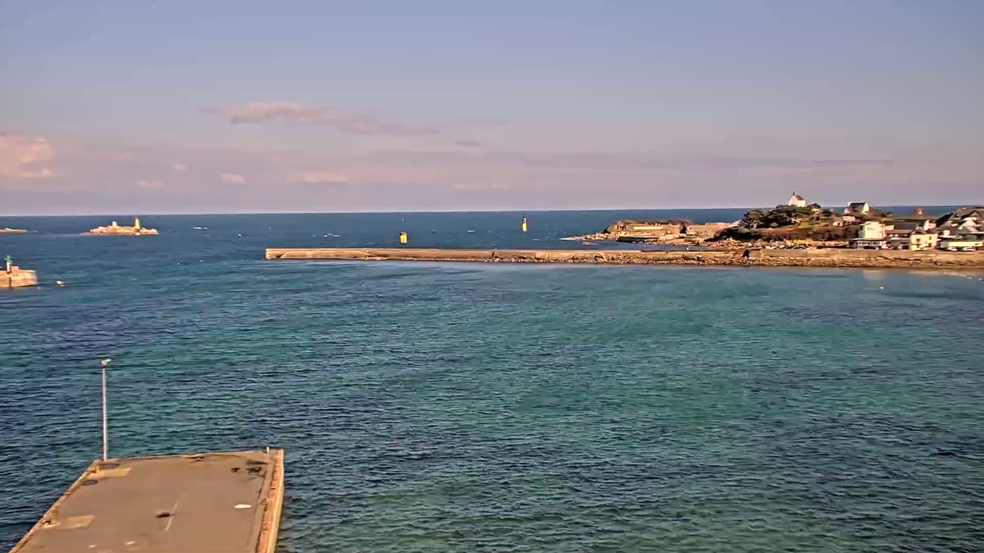 A calm, clear day shows a coastal scene with a small town nestled on a rocky point, a breakwater extending into teal-colored water, and a distant island.