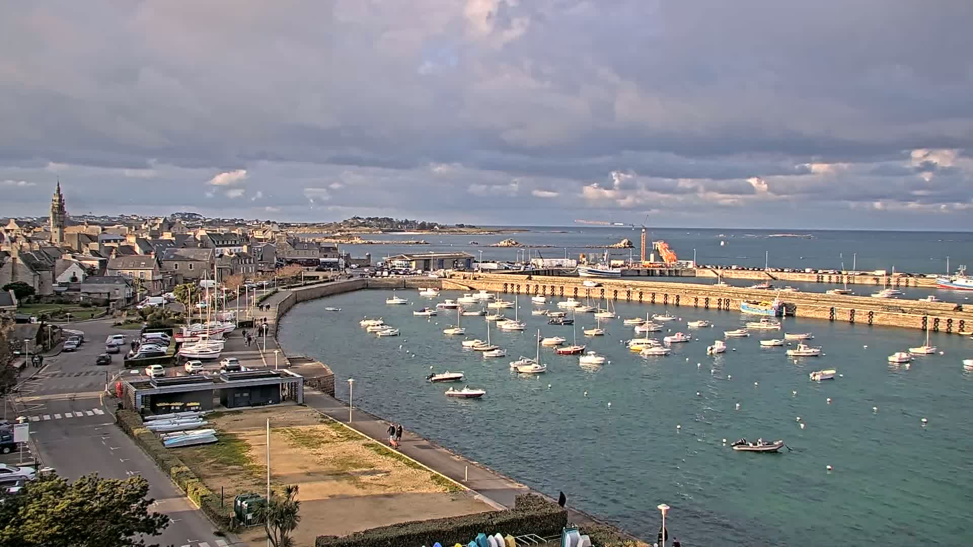 A harbor filled with numerous sailboats and small motorboats is situated next to a town under a partly cloudy sky.