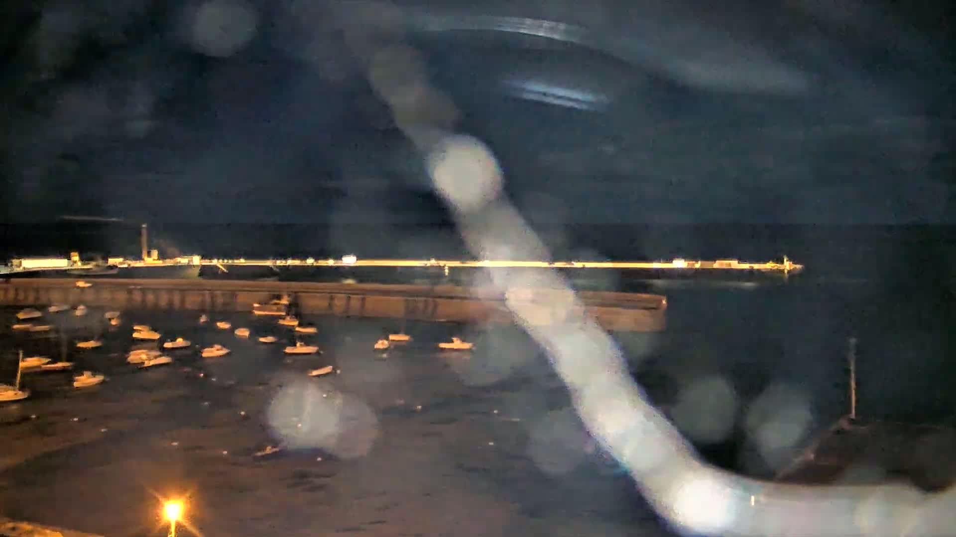 A nighttime view of a harbor filled with numerous small boats, seen through rain-streaked glass, under a dark, cloudy sky.