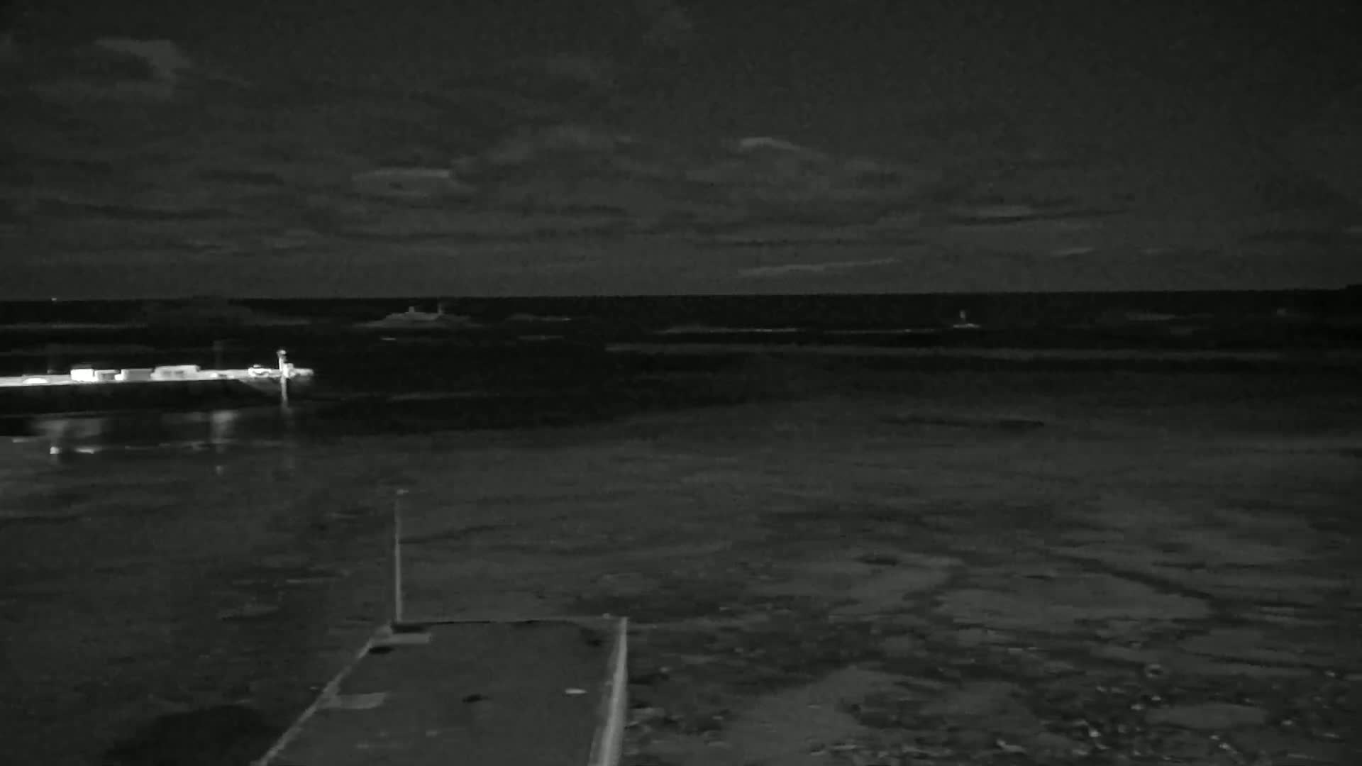 A nighttime view of a dark, calm body of water with a pier and distant lights under a cloudy sky.