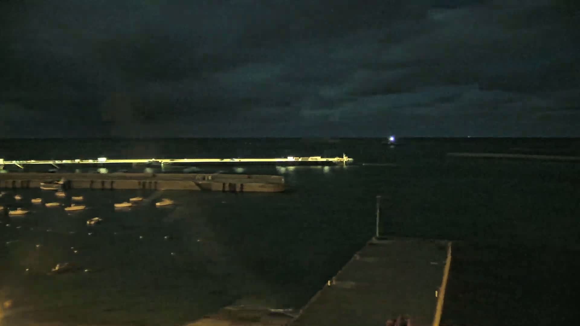 At night, under a dark, cloudy sky, a harbor shows several small boats moored near a long pier with a larger vessel beyond it.