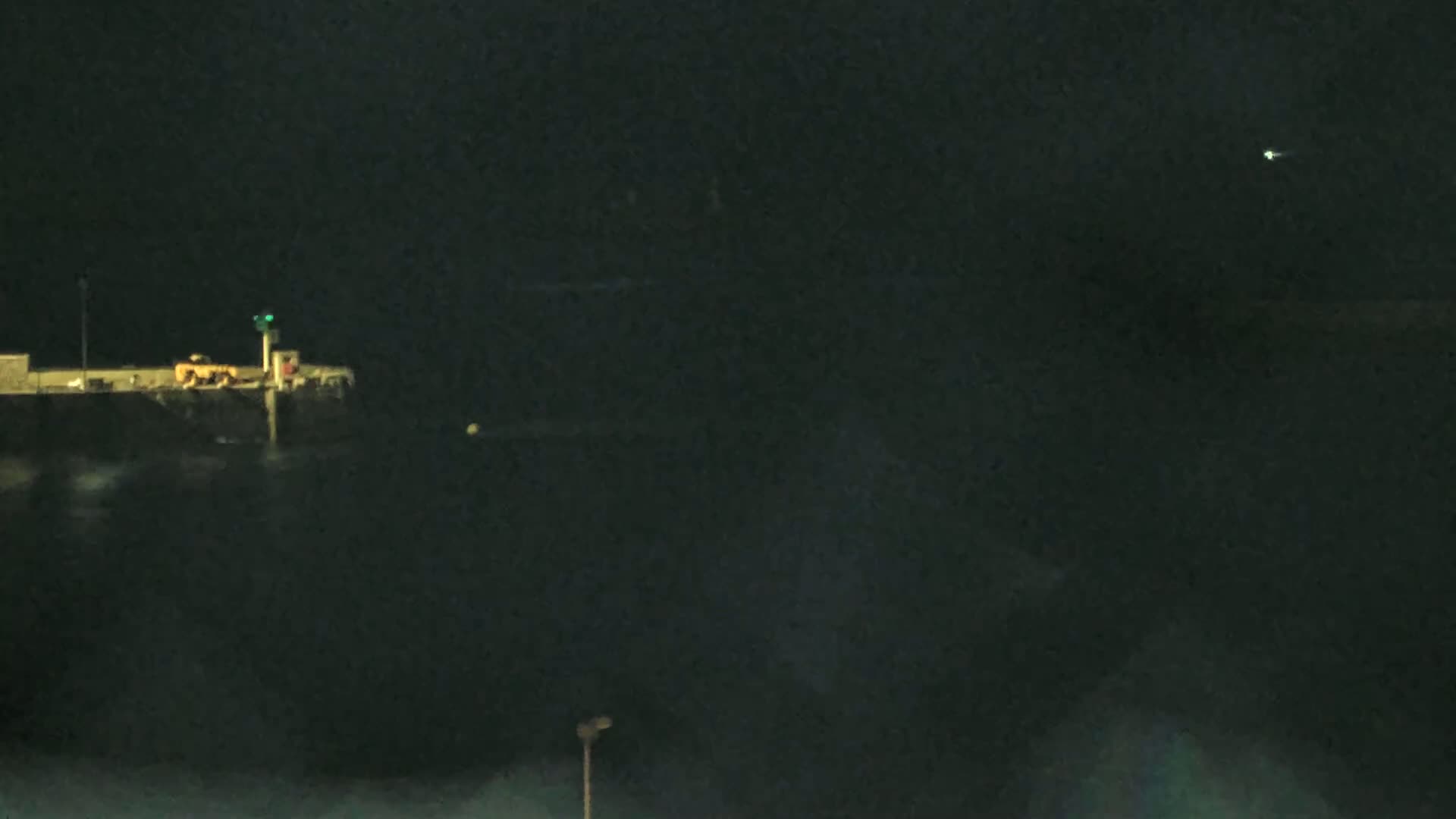 A dark, nighttime scene shows a pier with a light on it and a single light in the distance over dark water.