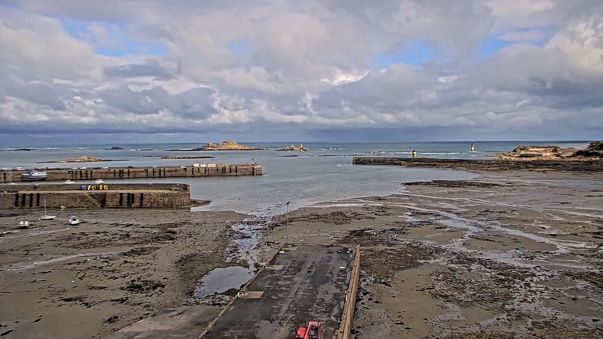 A harbor with stone walls is mostly exposed at low tide under a cloudy sky, with small boats and islands visible in the distance.
