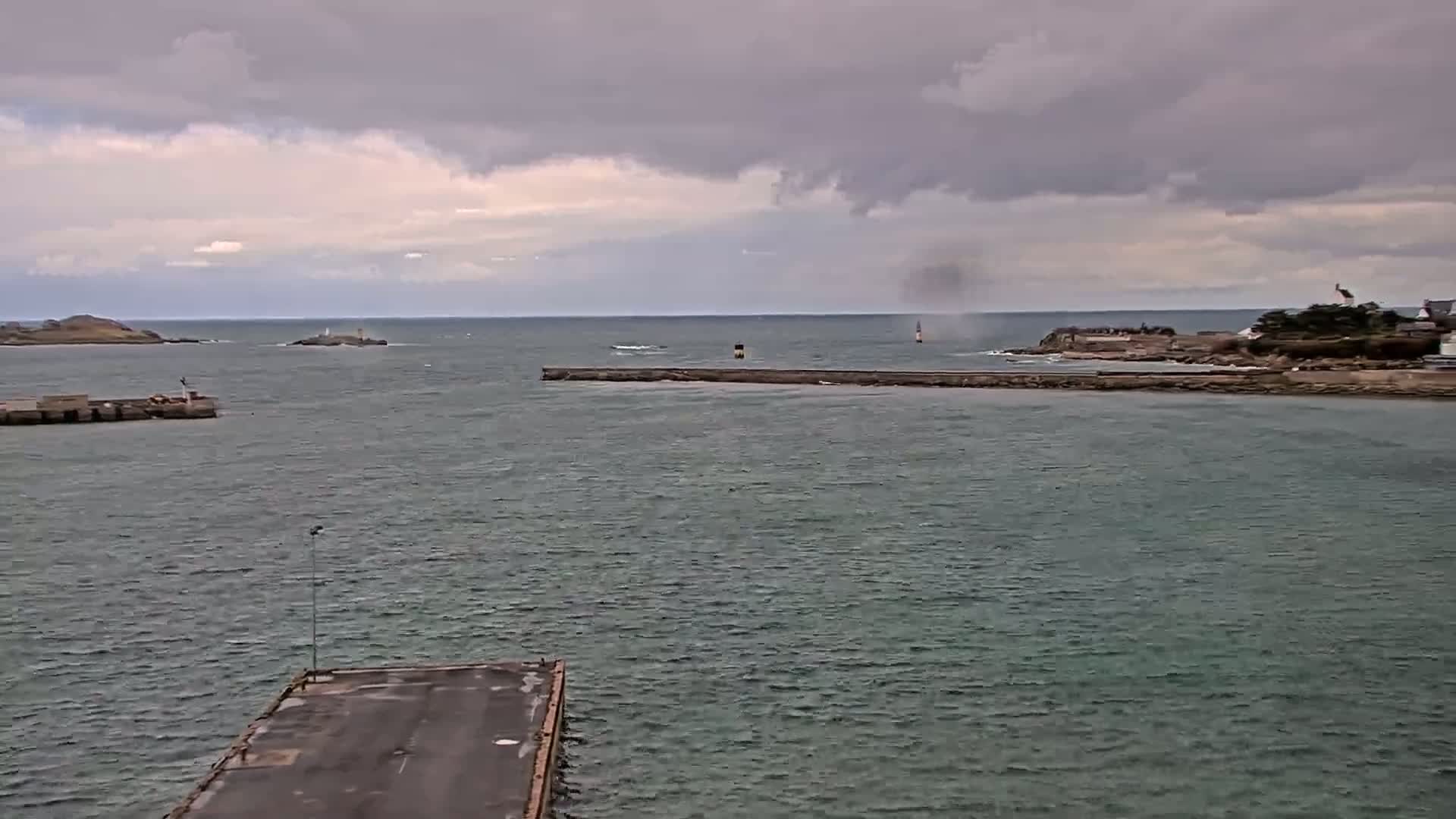A harbor scene under an overcast sky shows a calm body of water with a breakwater, small islands in the distance, and a long, empty dock in the foreground.