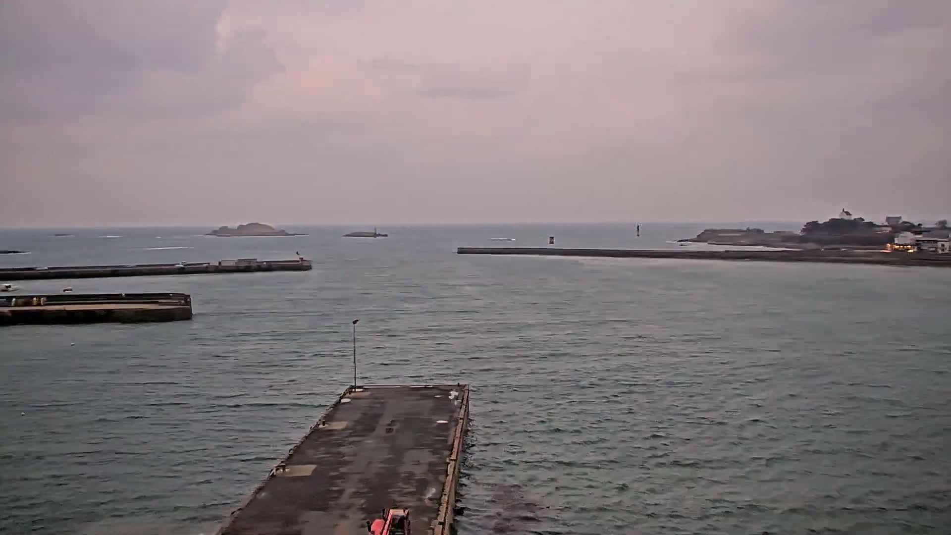 A long pier extends into a choppy, gray sea under an overcast sky, with distant landmasses and harbor structures visible.