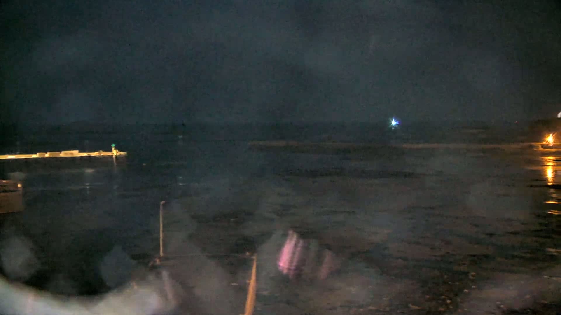 A dark, nighttime scene shows a body of water with a pier and faint lights in the distance under a cloudy sky.
