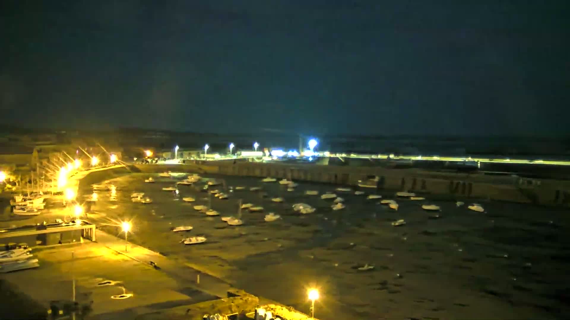 A nighttime harbor scene shows numerous small boats moored in calm water under a dark, clear sky, illuminated by streetlights along a paved walkway.