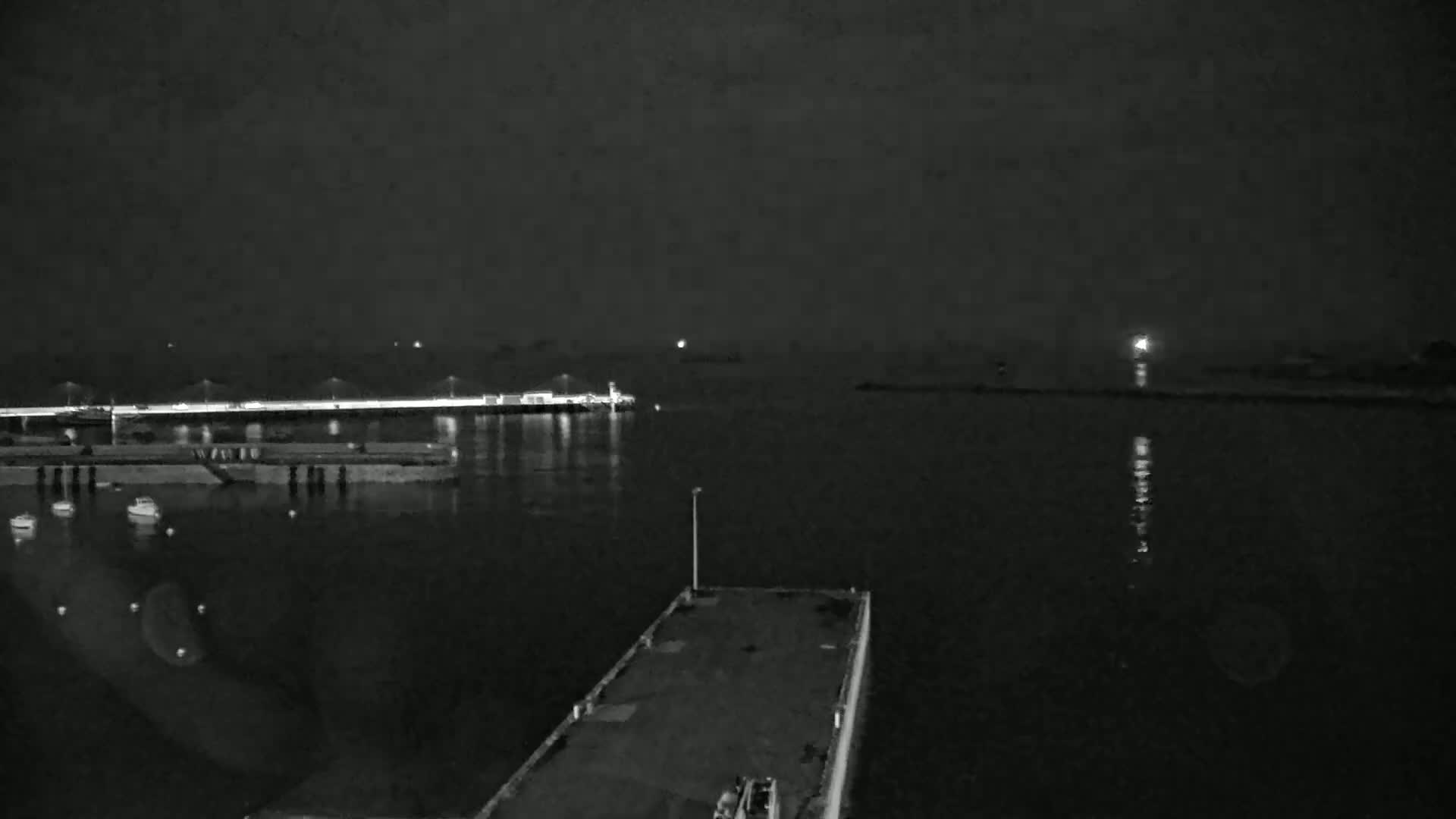 A nighttime view of a dark harbor with a few boats moored near a lit pier, under a hazy sky.