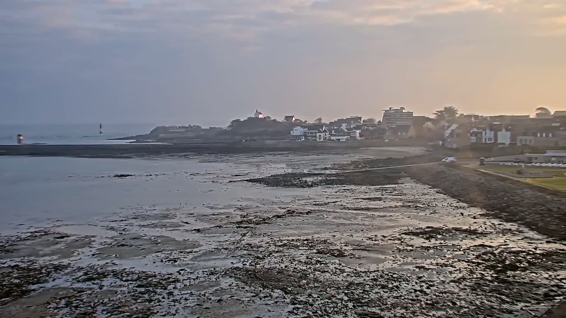 A coastal town is seen at low tide under a cloudy, dusky sky, with a lighthouse visible in the distance.