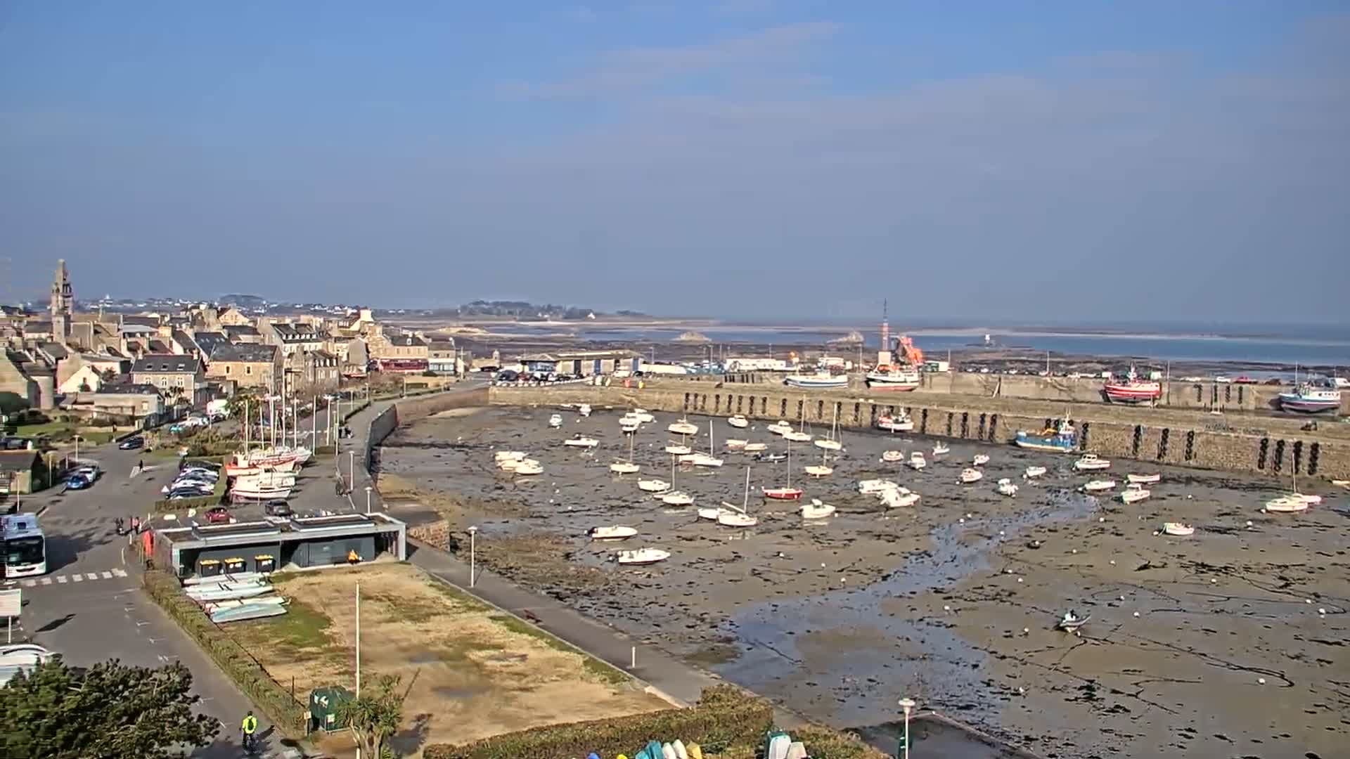 A harbor filled with numerous small boats sits mostly dry at low tide on a sunny day, adjacent to a town with buildings and parked cars.