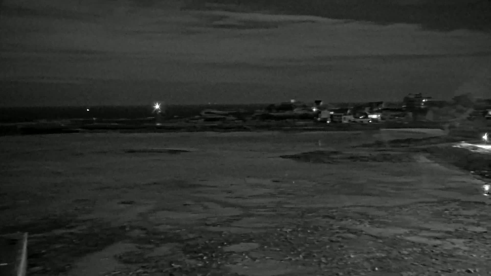 A dark, nighttime coastal scene shows a flat, wet expanse of land extending from a shoreline with a few distant lights visible under a mostly cloudy sky.