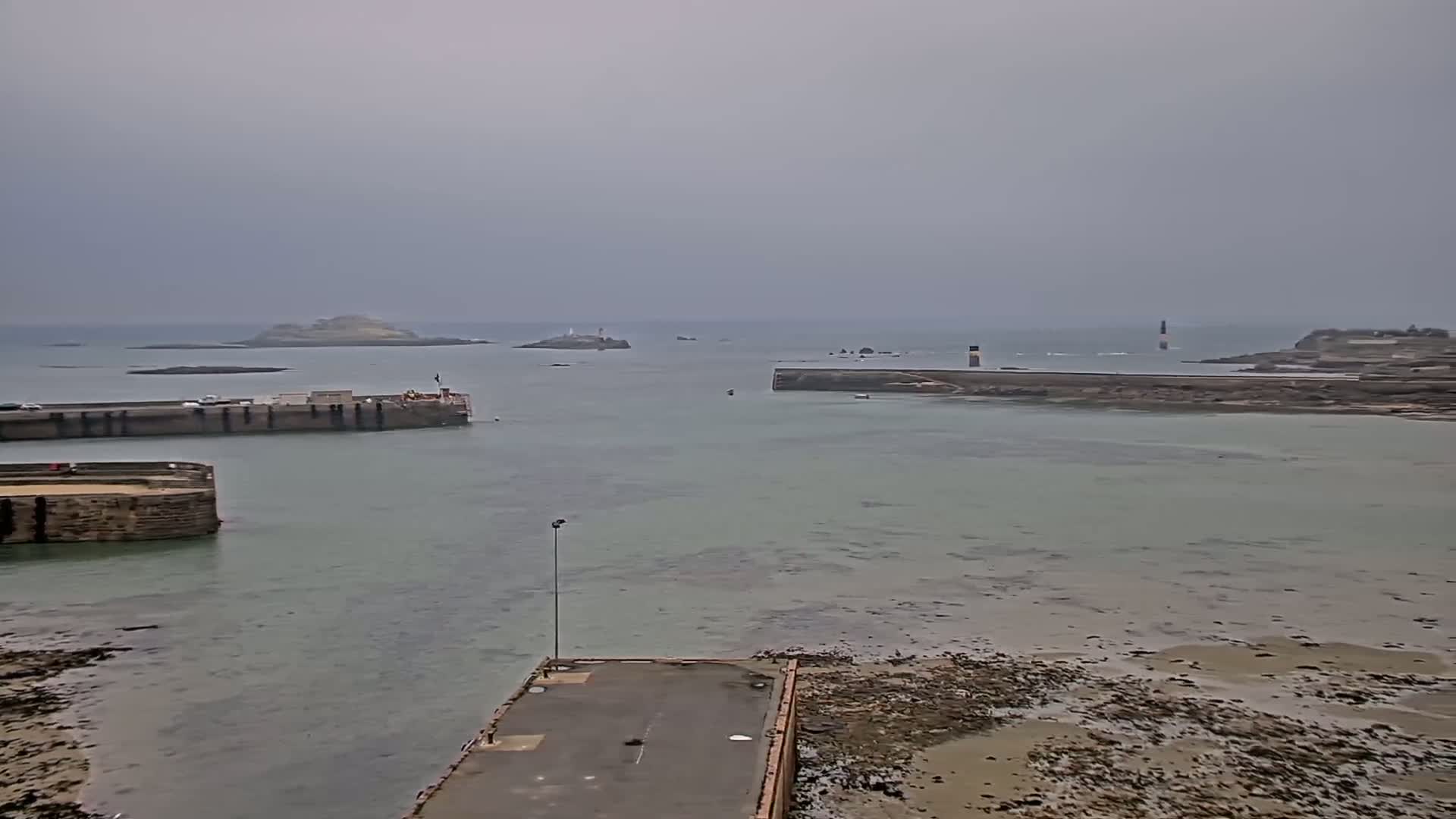 A harbor with calm, light-green water under an overcast sky, featuring stone harbor walls, a small pier, and distant islands with lighthouses.